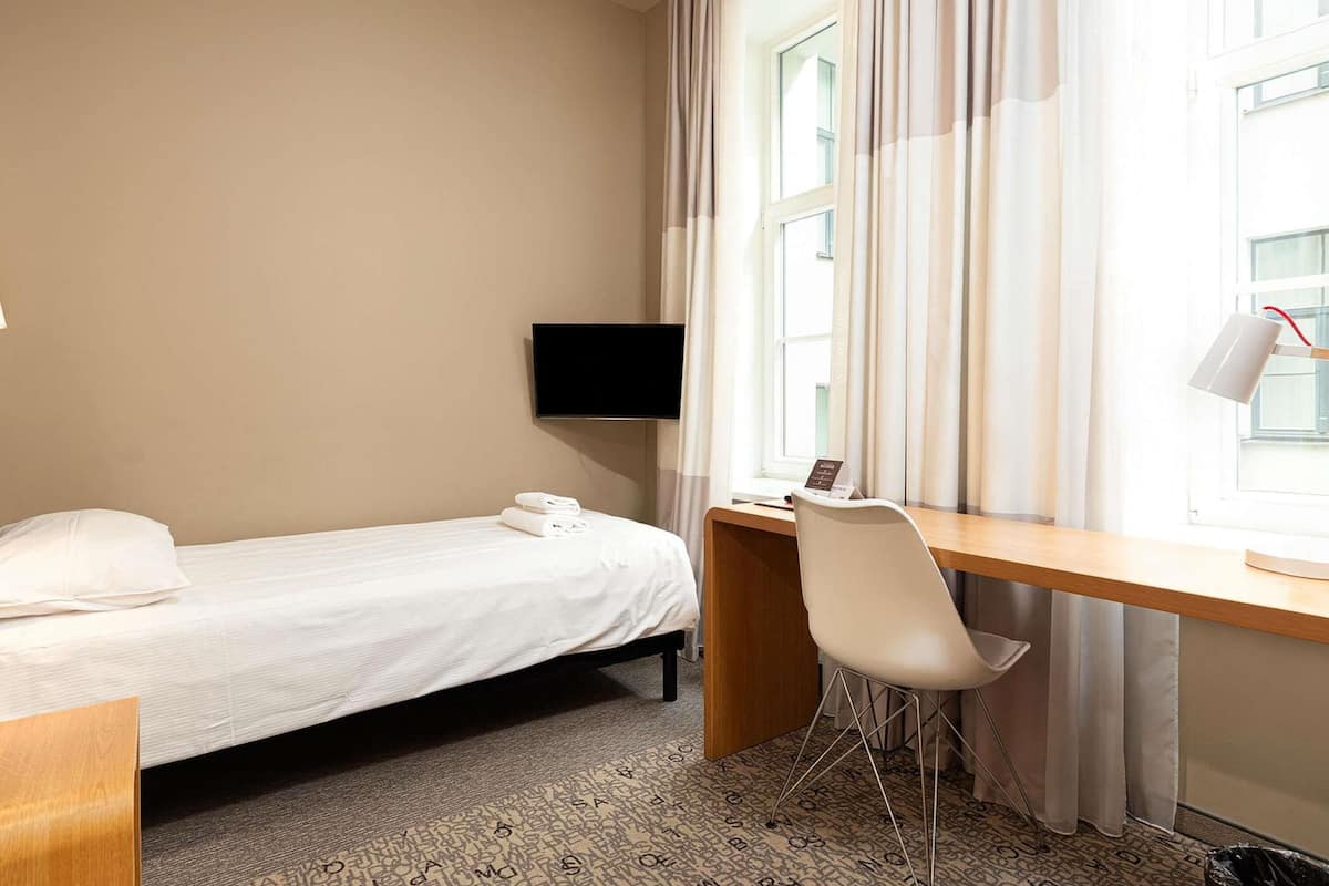Habitación de hotel con una cama individual, un escritorio con silla, televisor y ventana con cortinas.