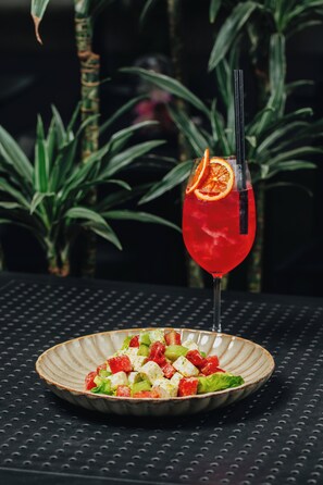 A cocktail in a glass with an orange slice garnish, a plate of cubed cheese salad, and a dark textured table surface. - ibis Riga Centre (Riga)