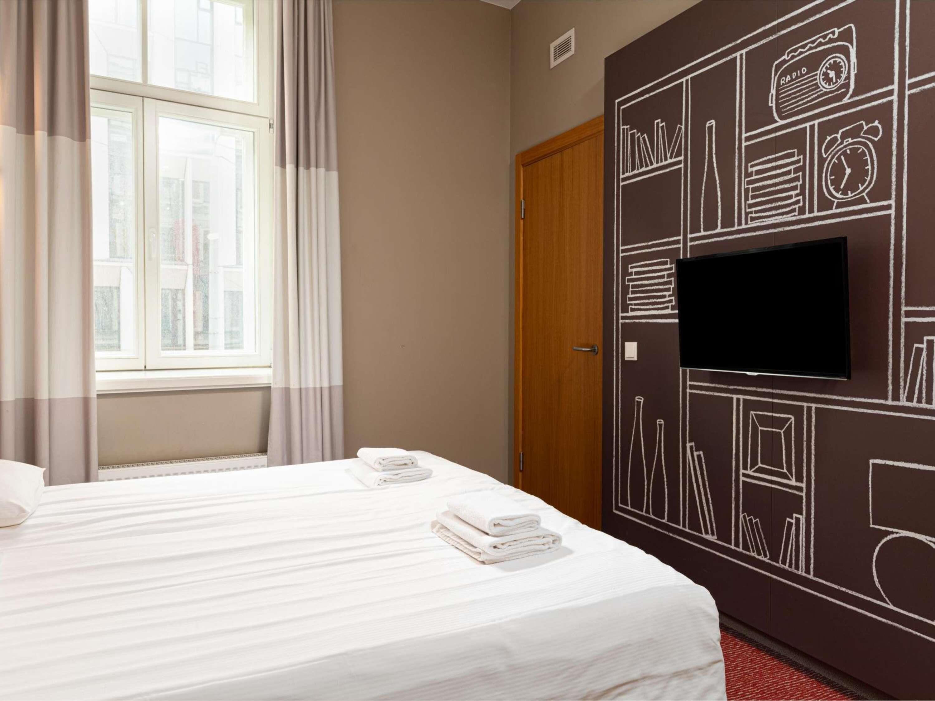 Ein Hotelzimmer mit einem Bett, einem Fernseher, einem Fenster mit Vorhängen sowie einer Wand mit Radio und einer Uhr-Design.