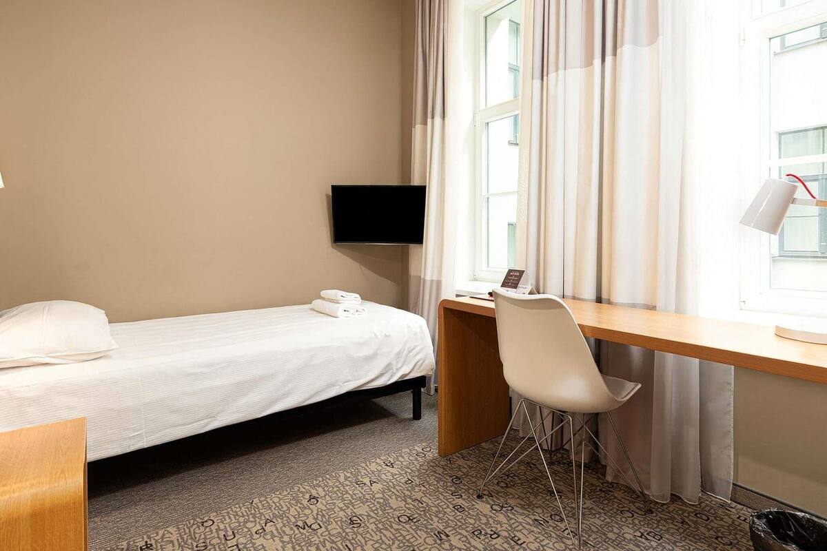 A hotel room with a bed, a desk with a chair, a TV, and a window with curtains.