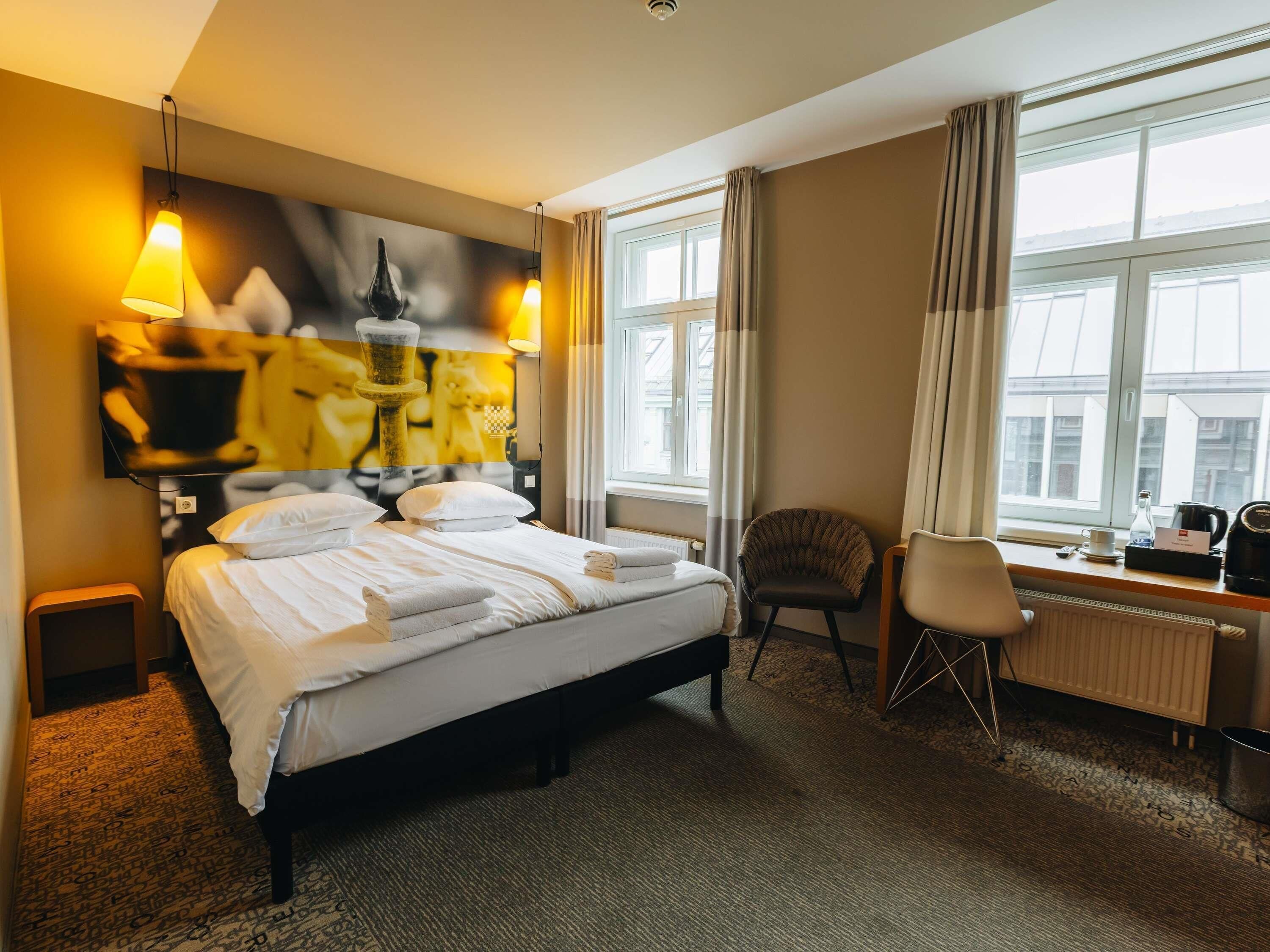Habitación de hotel con cama, escritorio, silla, ventana con cortinas y una obra de arte en la pared.