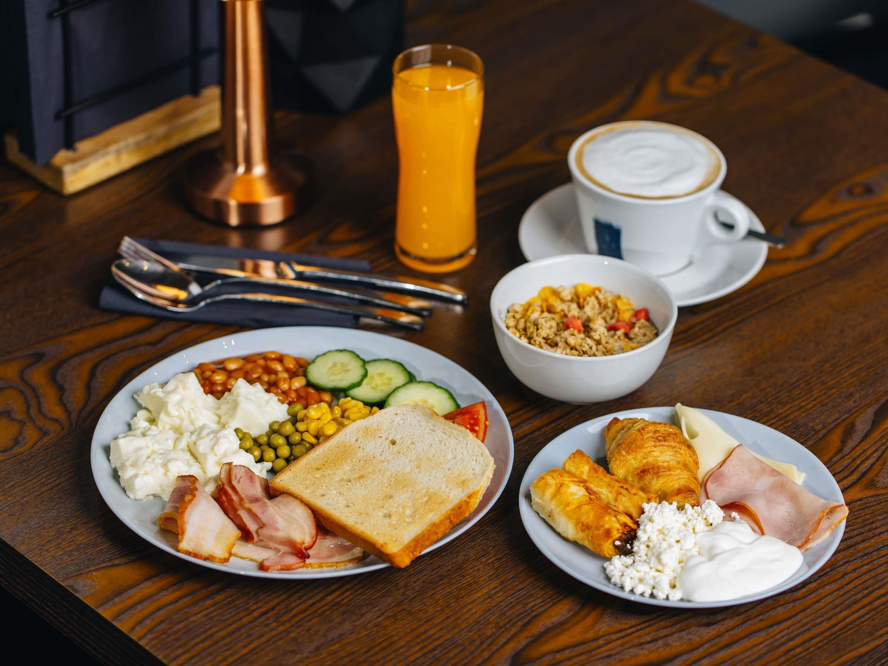 A breakfast setting with plates of eggs, toast, beans, and ham, a bowl of cereal, a glass of orange juice, and a cup of coffee on a wooden table.