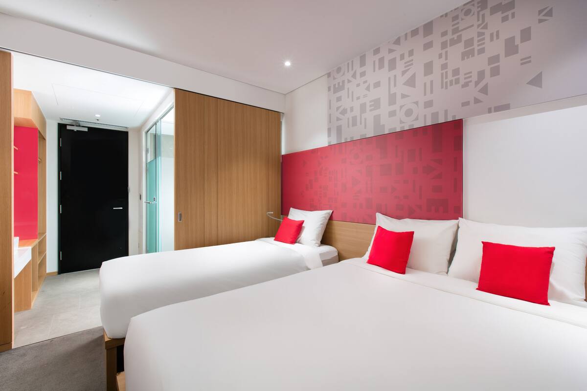 Une chambre d’hôtel moderne avec deux lits, une tête de lit en bois et un mur d’accent rouge.