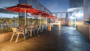 Un espace de restauration en plein air avec des tables et des chaises, des parasols rouges et une vue sur les toits d'une ville au crépuscule.