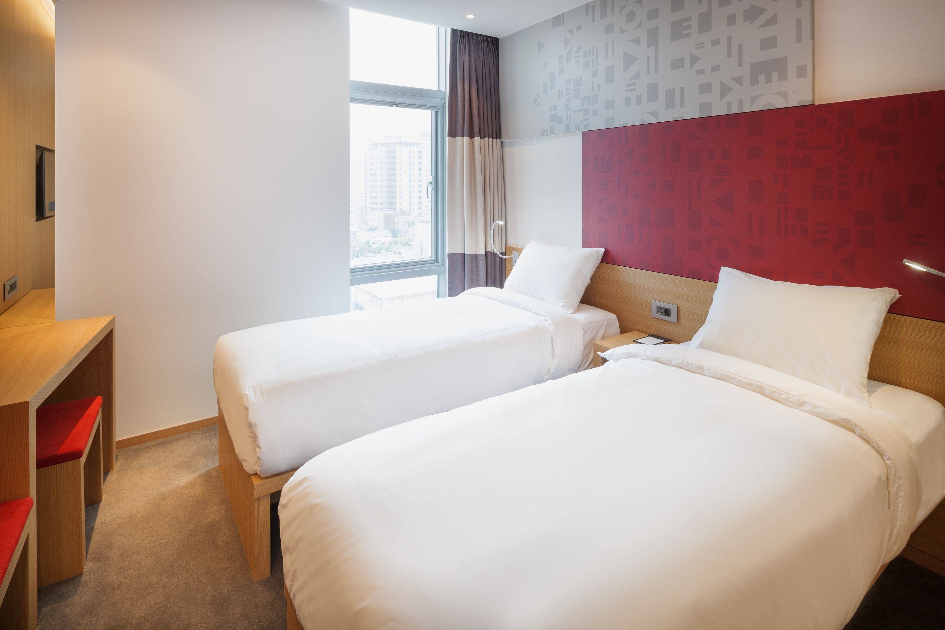 Habitación de hotel con dos camas individuales, un escritorio de madera con una silla roja, una ventana con cortinas y una pared de acento roja.
