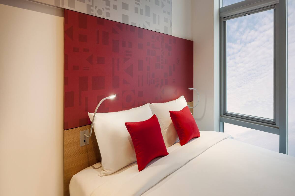 Une chambre d’hôtel moderne avec un grand lit, des oreillers rouges et blancs, une tête de lit ornée de motifs géométriques et une fenêtre donnant sur le ciel.
