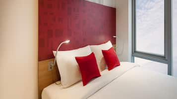 A modern hotel room with a large bed, red and white pillows, a headboard with geometric patterns, and a window with a view of the sky.