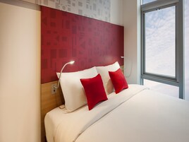 Una habitación de hotel moderna con una cama grande, almohadas rojas y blancas, un cabecero con diseños geométricos y un ventanal con vistas al cielo.
