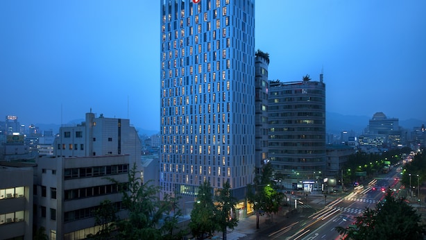 Un hôtel moderne de grande hauteur, reconnaissable à son logo Travelodge bien visible, entouré de bâtiments urbains et bordé par une rue animée, le tout baigné par la lumière du crépuscule.