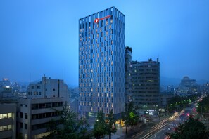 Ett modernt höghotell med en tydlig Travelodge-logotyp, omgivet av byggnader i stadsmiljö och en livlig gata i skymningen.