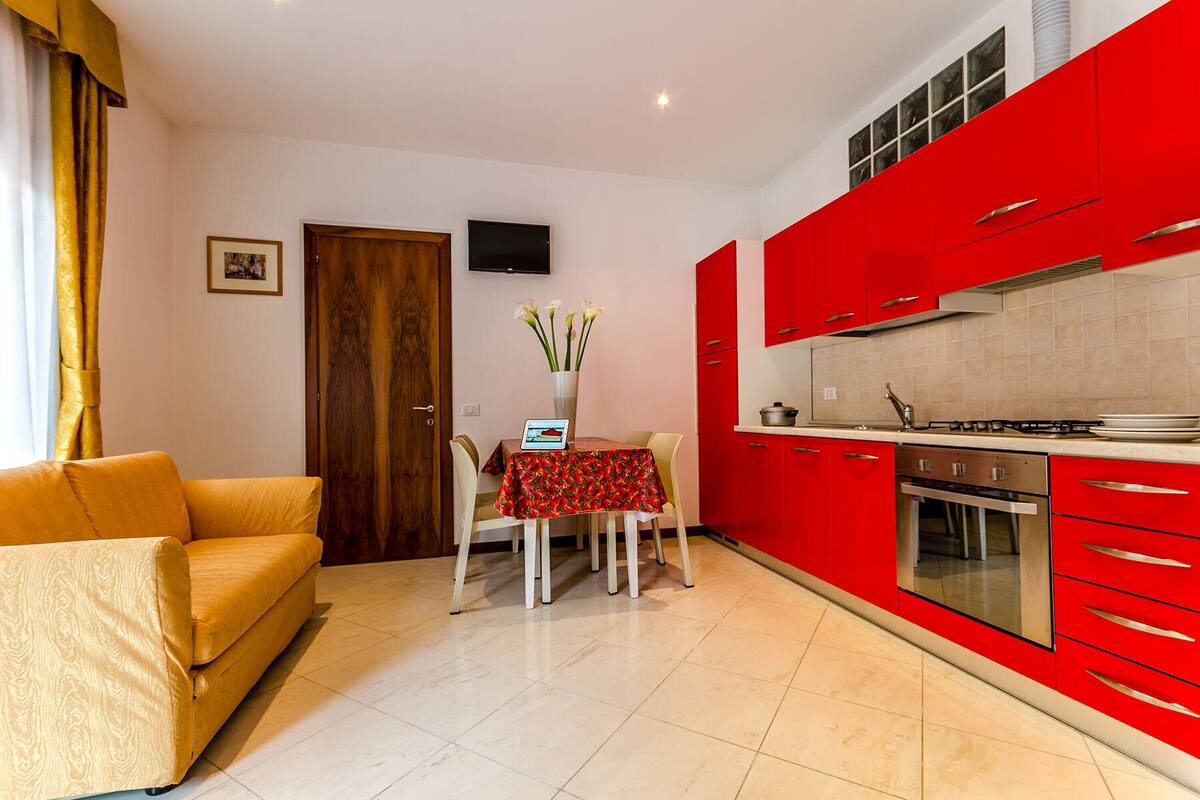 A modern living room with a sofa and a dining table with a red tablecloth. There is a kitchen with red cabinets and a stove.