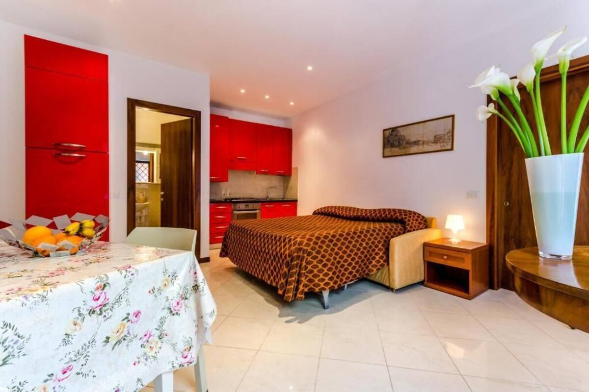 A compact hotel room with a bed, bedside table, and a small table with fruit. A kitchen with red cabinets is visible through an open door.