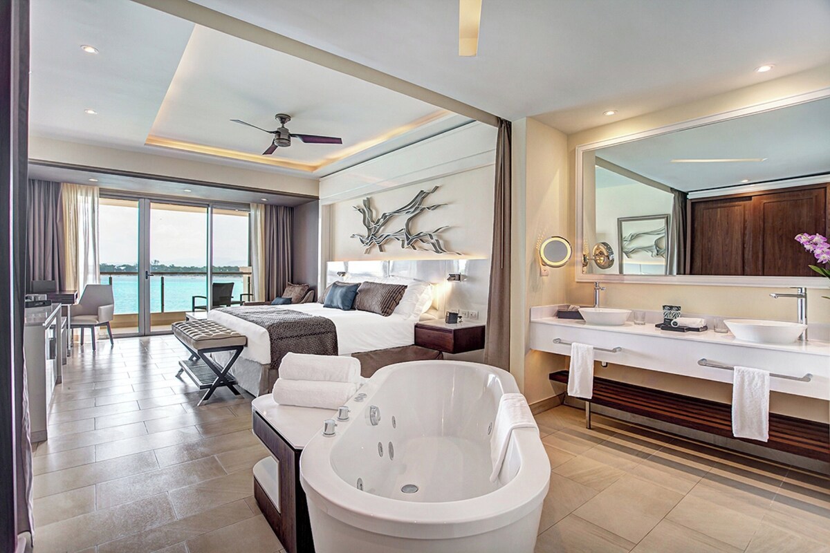 A modern hotel room with a large bed, a sitting area with a view, a large bathtub, and a bathroom with a vanity and mirror.