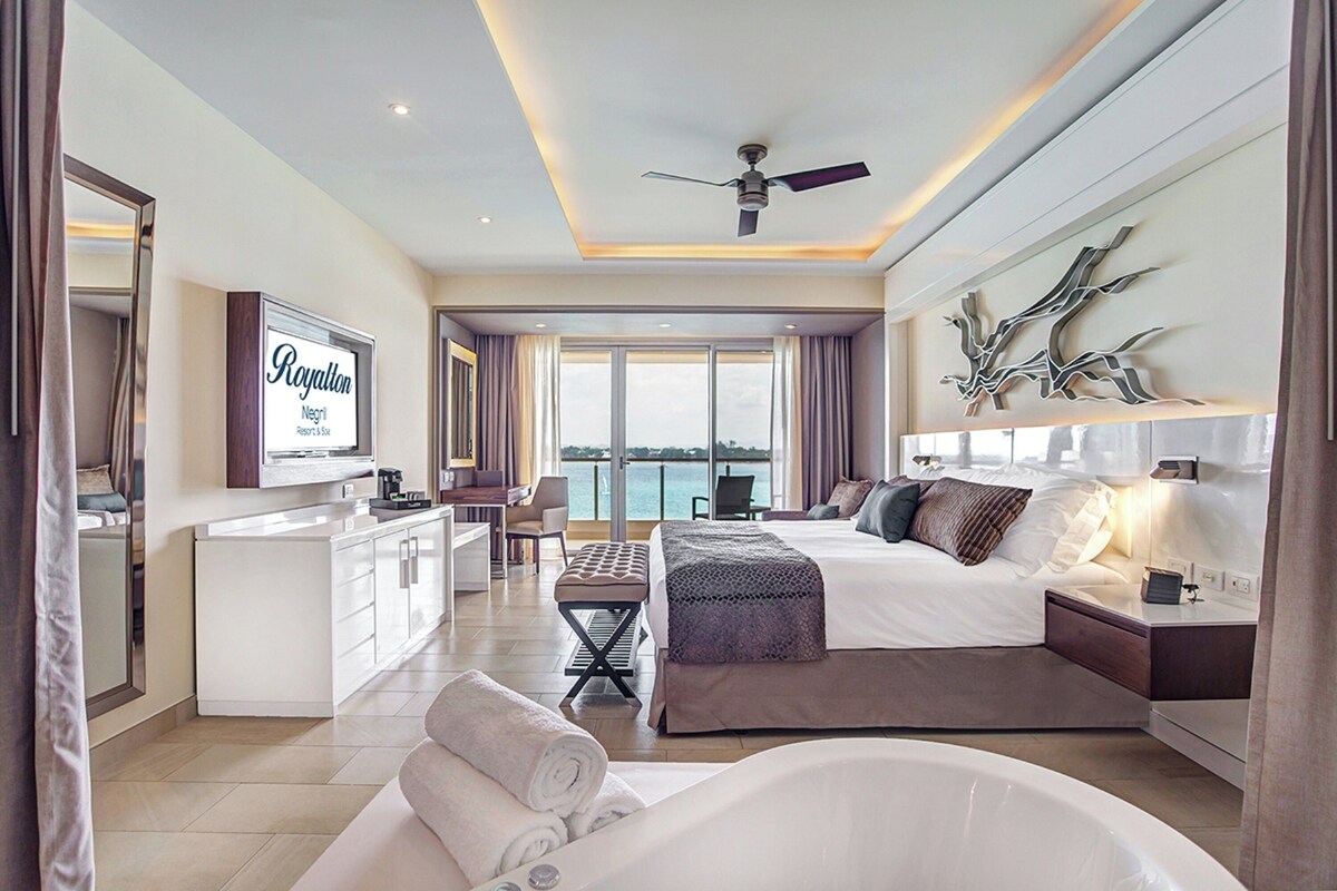 A modern hotel room with a king-sized bed, a sitting area with a desk, a large window with a view, a ceiling fan, and a mounted television.