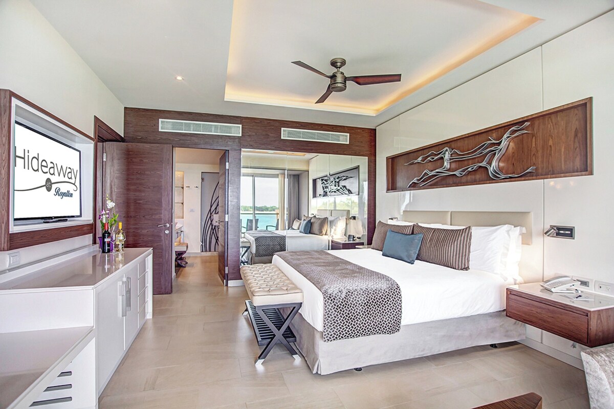 A modern hotel room with a large bed, a desk, a ceiling fan, and a view of the sea.