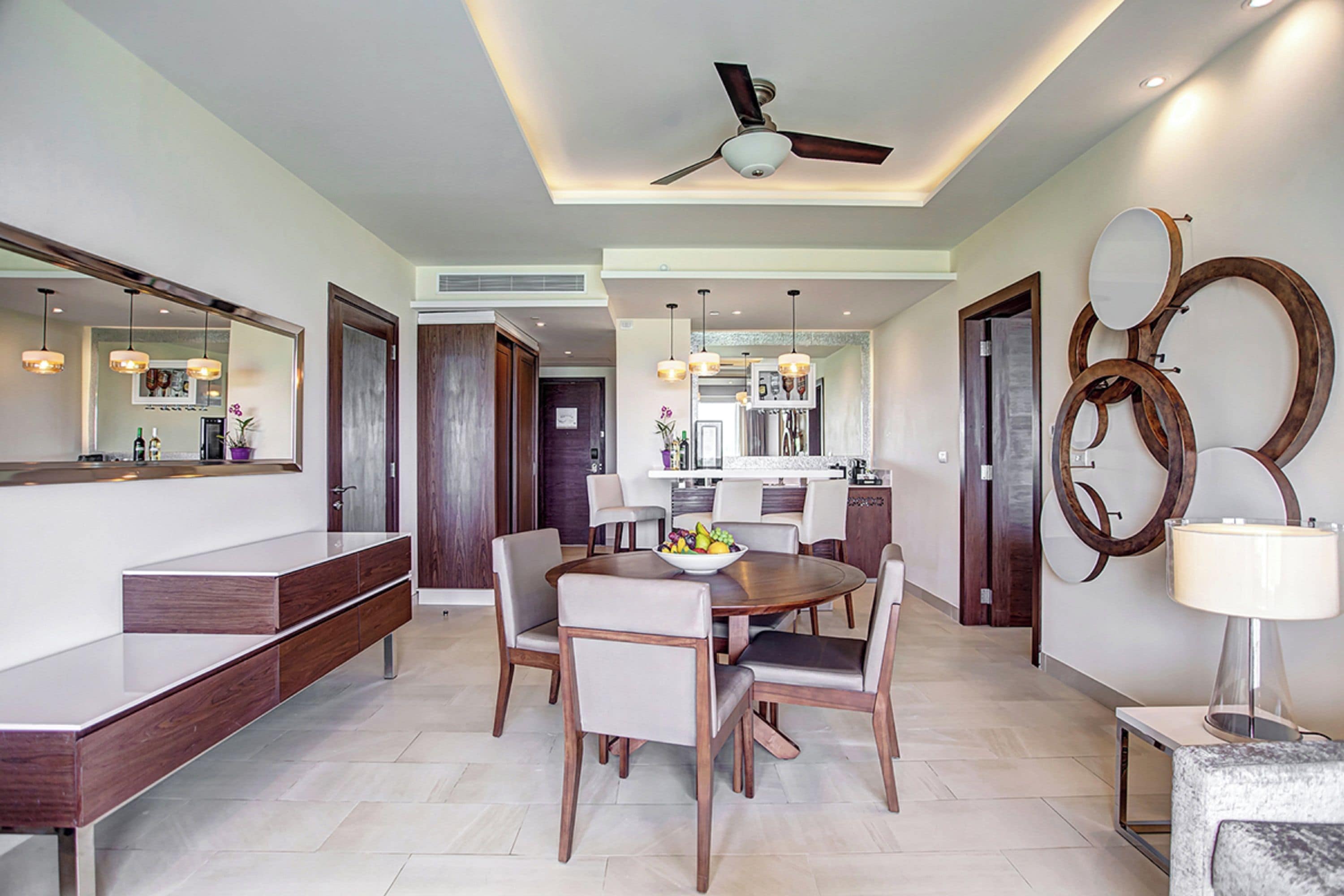 A modern dining area with a round table, chairs, and a bowl of fruit. There is a ceiling fan, pendant lights, and a mirror on the wall.