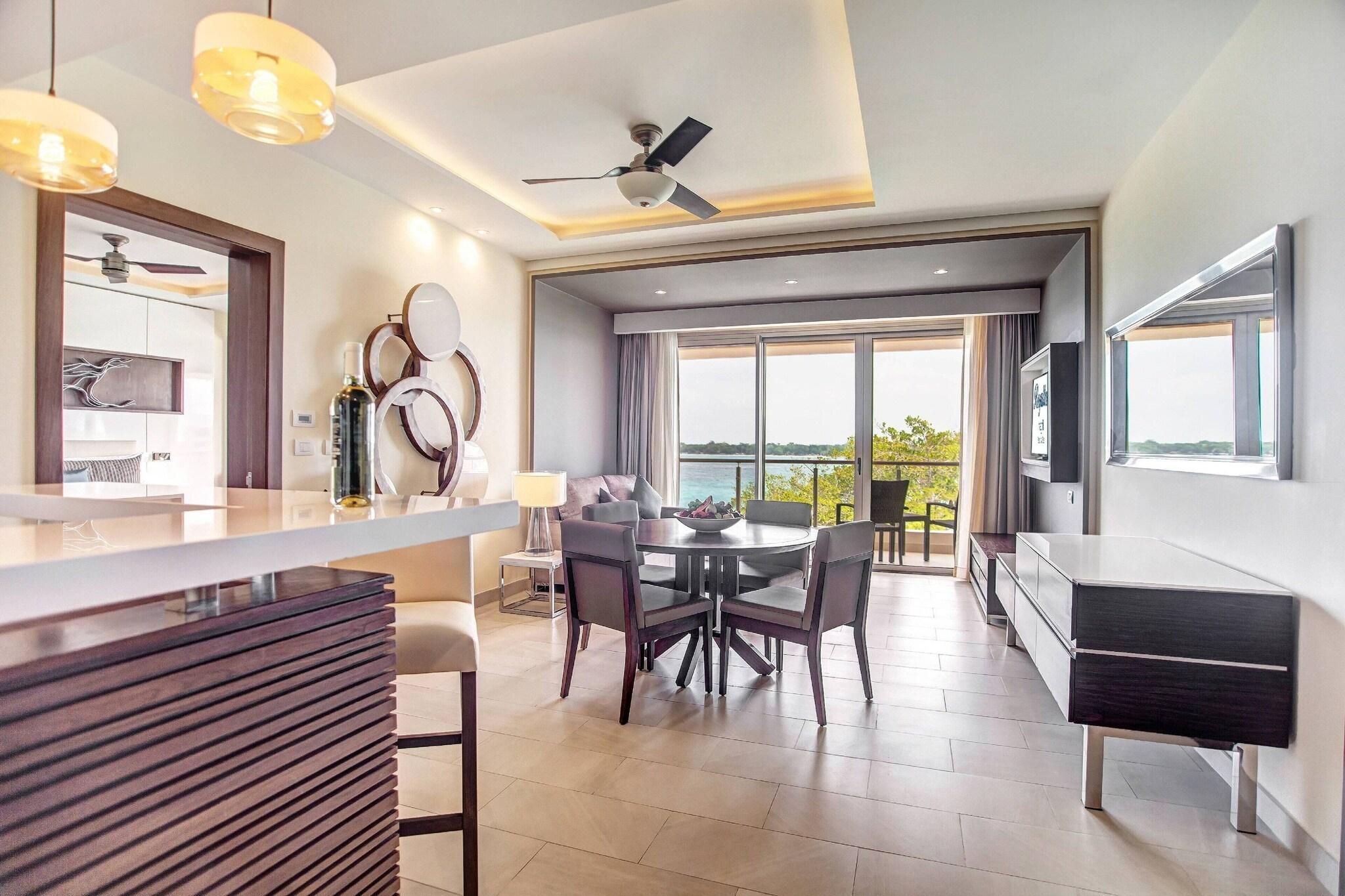A modern hotel room with a dining area, a large window with a view, a ceiling fan, and a TV mounted on the wall.
