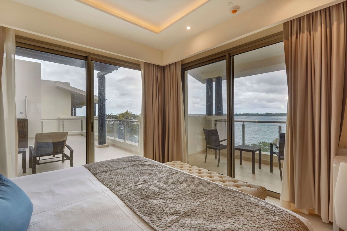 A modern bedroom with a large bed, a view of the sea, and a balcony with seating.