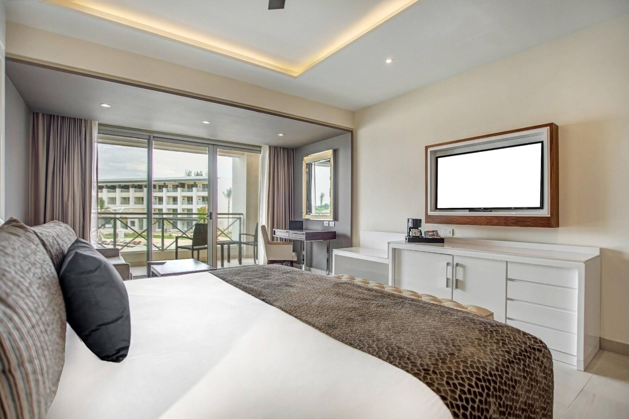 A hotel room with a large bed, a desk, a chair, a TV, and a balcony with a view of buildings.