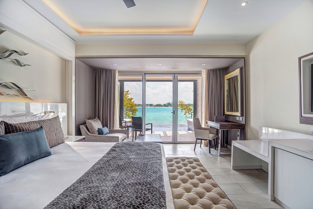 A modern hotel room with a large bed, a desk, and a view of the sea.