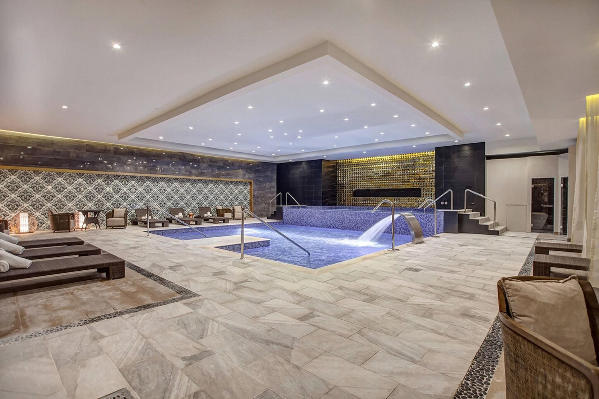 A spacious indoor pool area with a hot tub, seating, and a staircase leading to an upper level.