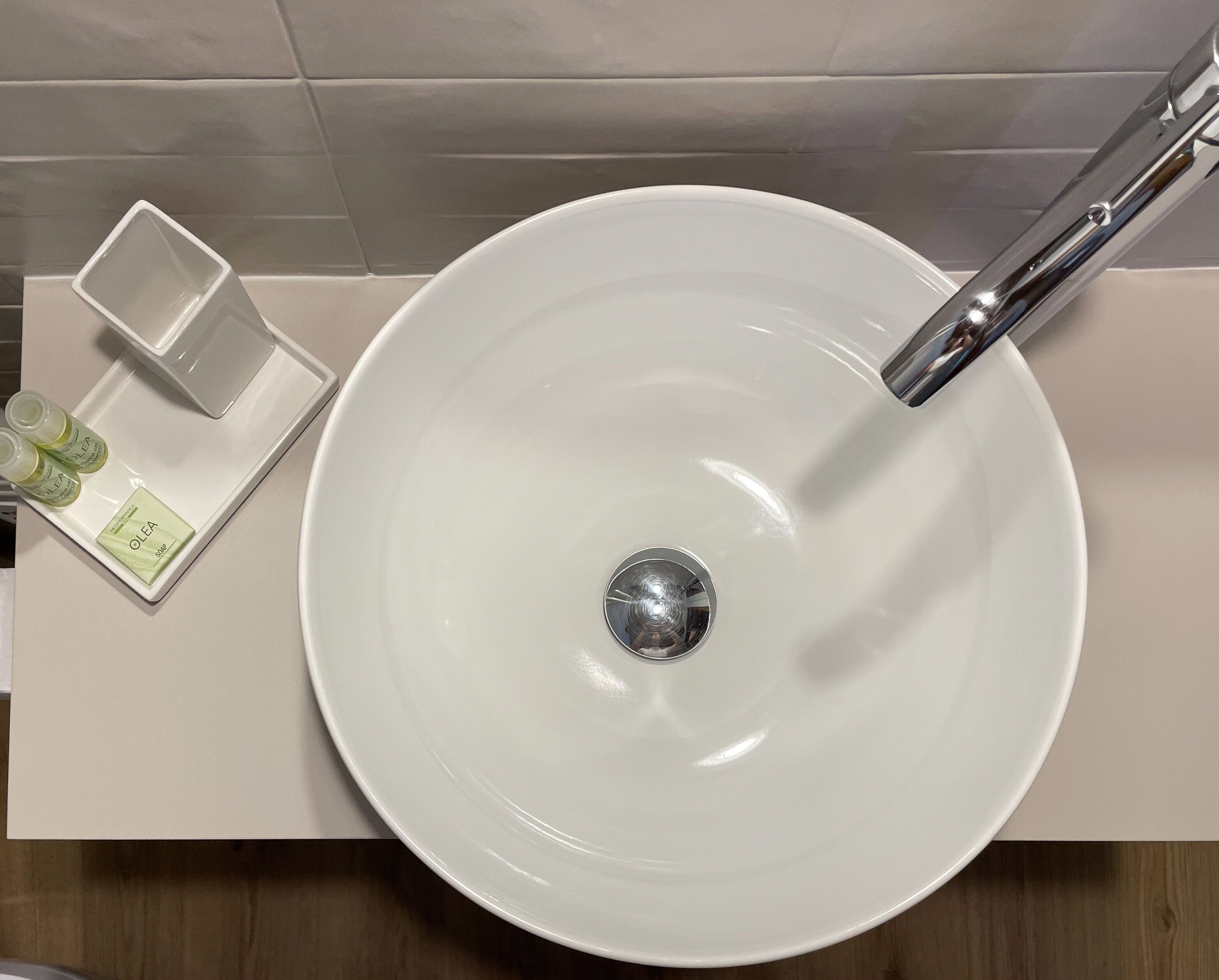 A white sink with a chrome faucet, a small rectangular container with a bottle and a bar of soap, and a wooden surface beneath.