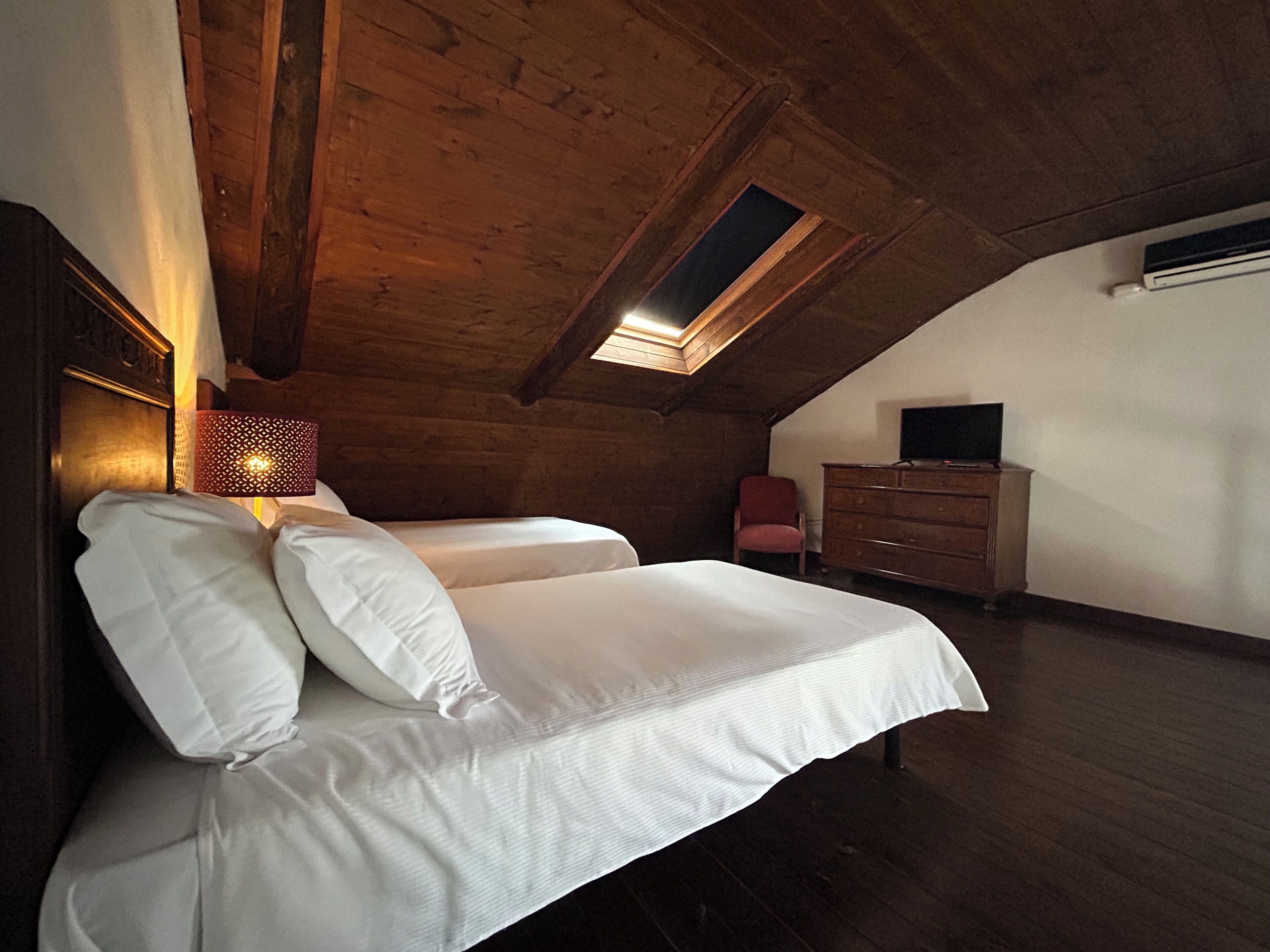 A bedroom with a wooden ceiling, a skylight, a bed with white linens, a dresser, and a television.