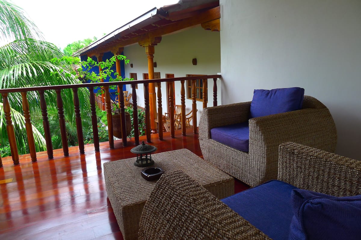 A balcony with wicker furniture, a small table with a decorative item, and a view of lush greenery and a building in the background.