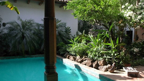 Una piscina con agua azul cristalina, rodeada de plantas tropicales y un pilar de piedra.
