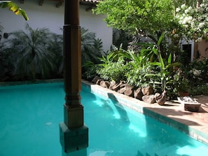 A swimming pool with clear blue water, surrounded by tropical plants and a stone pillar.