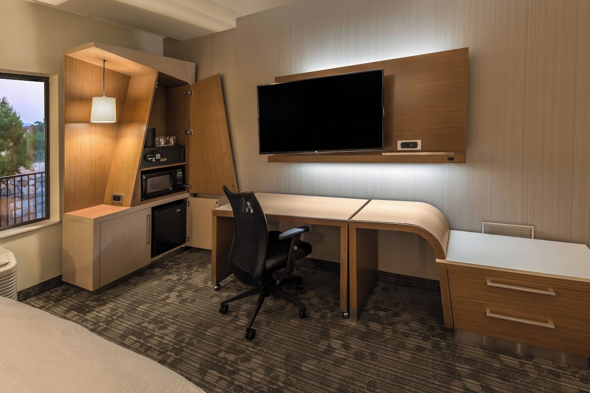 Quarto de hotel com escrivaninha, cadeira, televisão, micro-ondas e pequena cozinha.