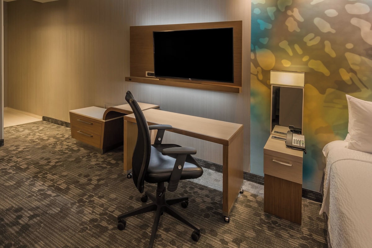 Quarto de hotel com escrivaninha, cadeira, cama e televisão presa à parede.