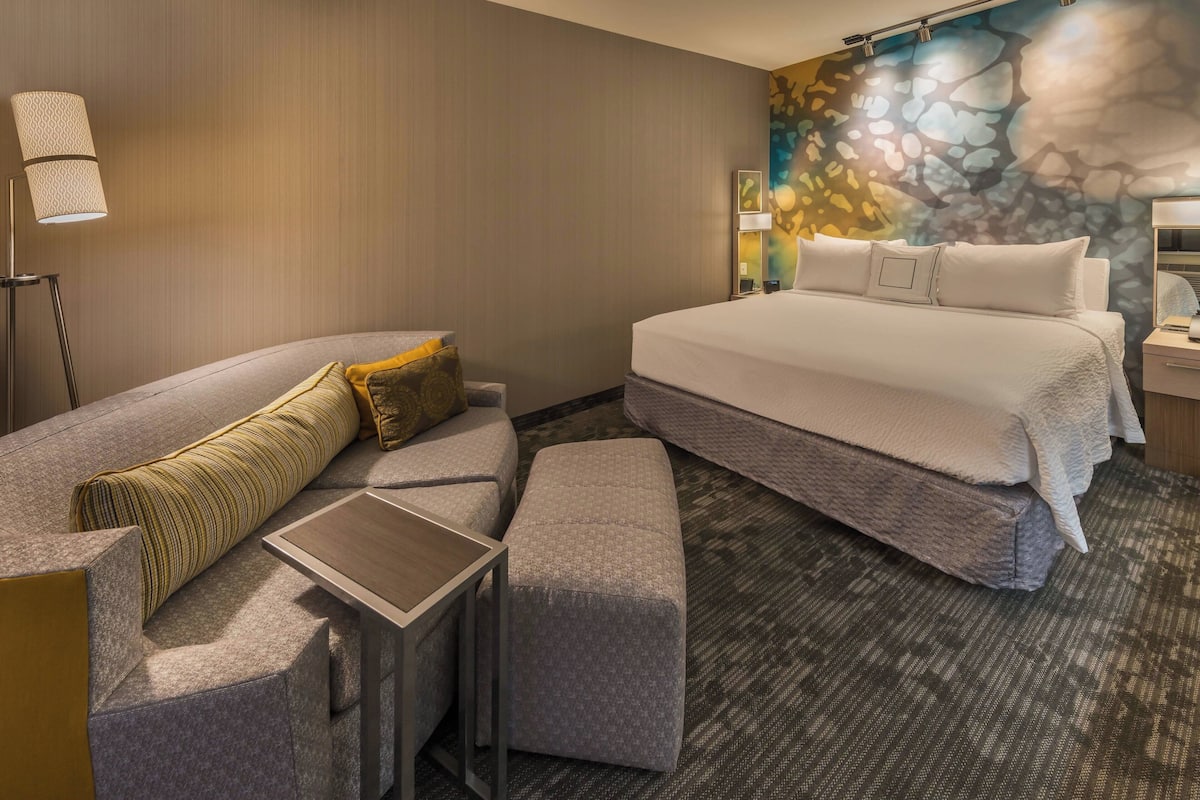 Quarto de hotel moderno com uma cama grande, um sofá, uma mesinha e chão coberto de carpete.