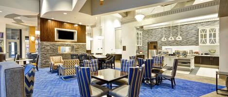A spacious hotel lobby with a central dining area, a kitchen, and a TV mounted on the wall.