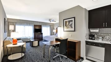 A hotel room with a bed, a desk, a chair, a small table with a lamp, a kitchenette, and a ceiling fan.