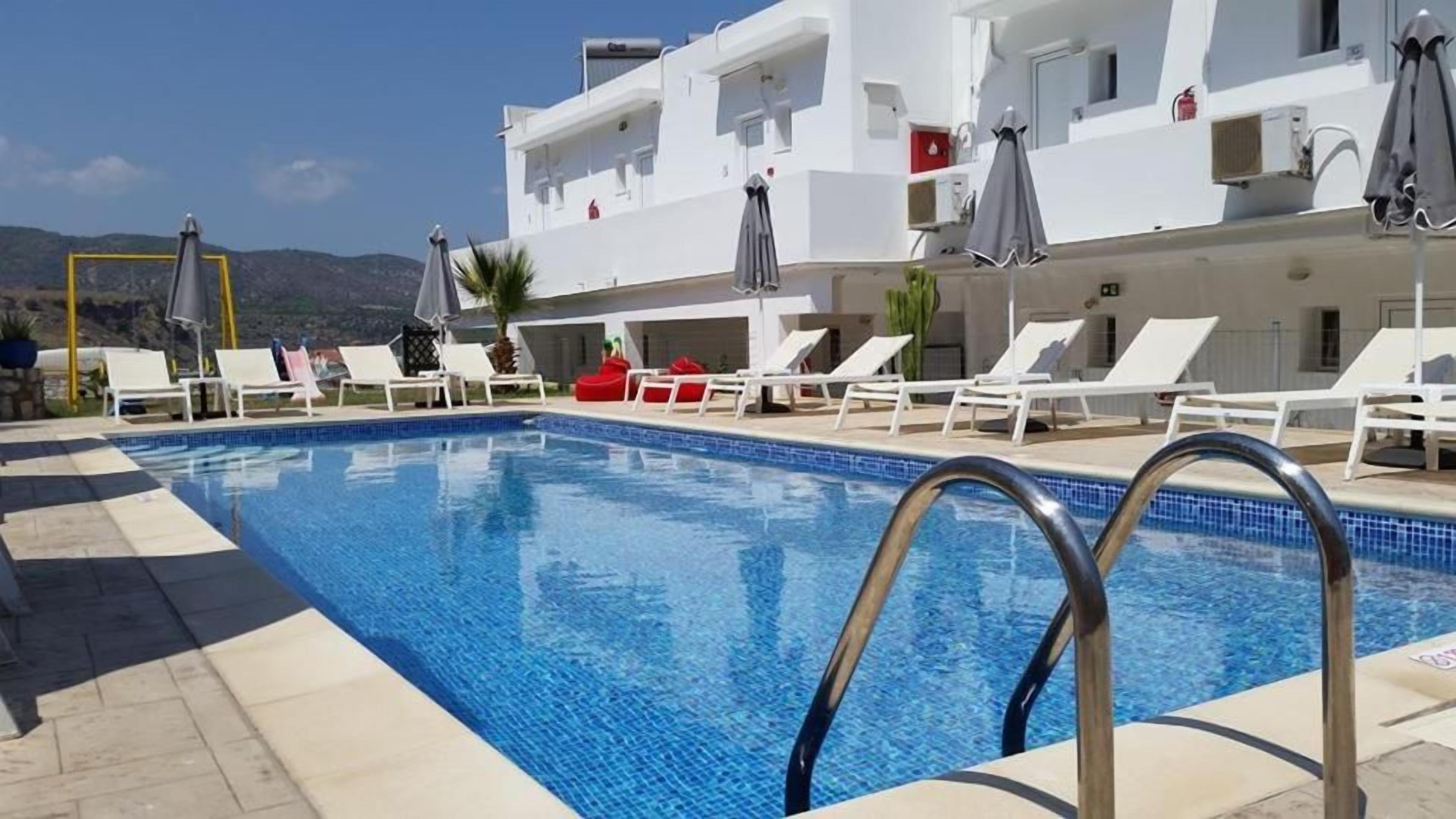 A swimming pool with sun loungers and umbrellas, surrounded by a white building with balconies.