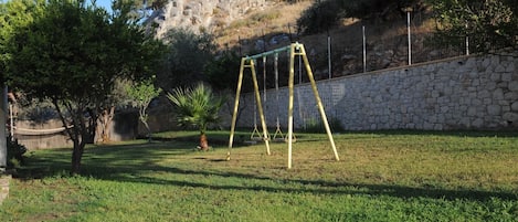 A grassy backyard with a swing set, a stone wall, and a tree.