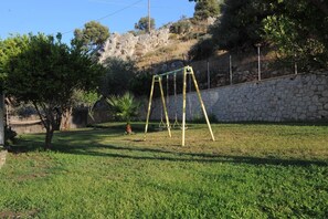 A grassy backyard with a swing set, a stone wall, and a tree. - Golden View Studios (Rhodes)