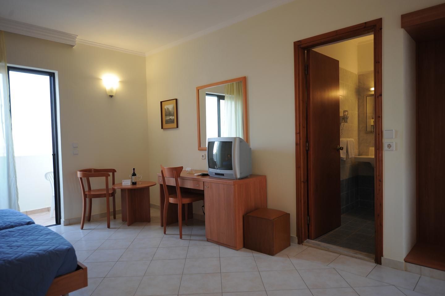 A hotel room with a bed, a desk with a television, a chair, a small table, and a door leading to a bathroom.
