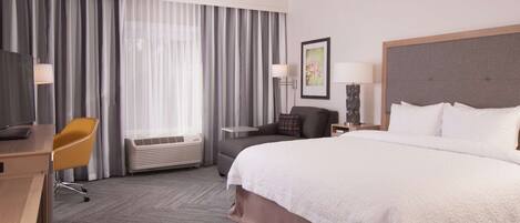 A hotel room with a large bed, a desk with a chair, a sofa, a TV, and a window with curtains.