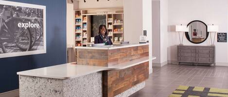Un'area reception di un hotel con un banco informazioni in legno, una targa a parete con la scritta 'welcome to augusta' e uno specchio sopra una consolle grigia.