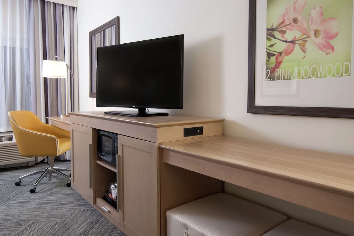 Habitación de hotel con una televisión de pantalla plana sobre un escritorio de madera, una silla amarilla, un cuadro enmarco con flores rosadas y una ventana con cortinas.