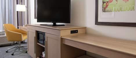 A hotel room with a flat-screen TV on a wooden desk, a yellow chair, a framed picture of pink flowers, and a window with curtains.