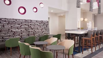 A modern hotel lobby with a dining area featuring round tables and chairs, a wall with text, and decorative lighting.