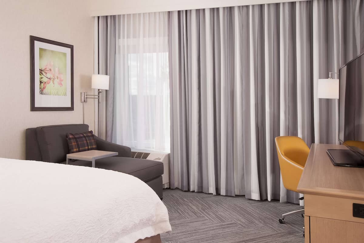 Habitación de hotel con cama, escritorio, silla, mesita, lámpara y ventana grande con cortinas.