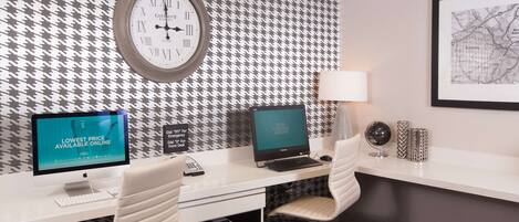 A hotel room with a houndstooth patterned wall, a round wall clock, a desk with two monitors, a printer, and a chair.