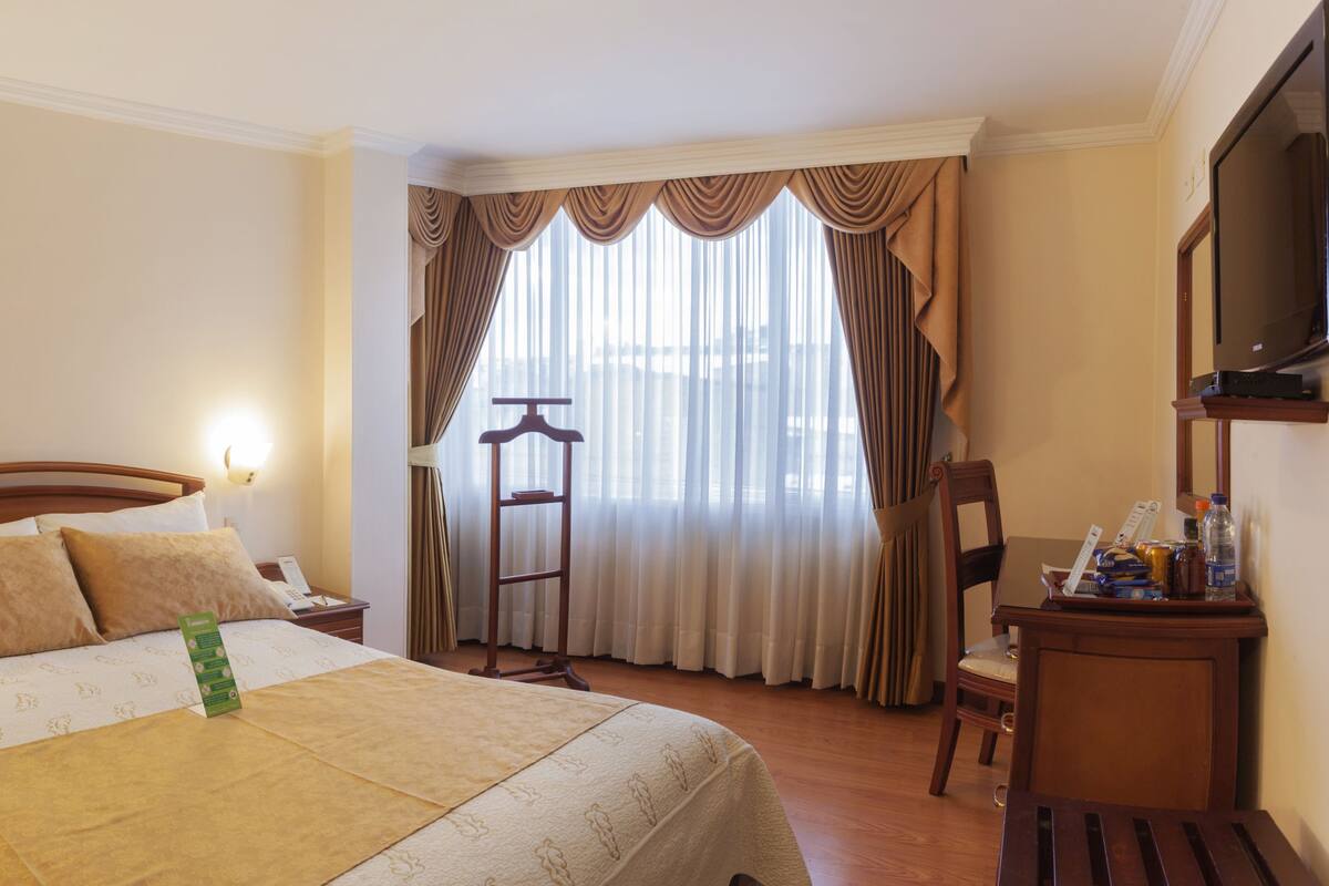A hotel room with a bed, bedside tables, a desk, a chair, a TV, and a window with curtains.