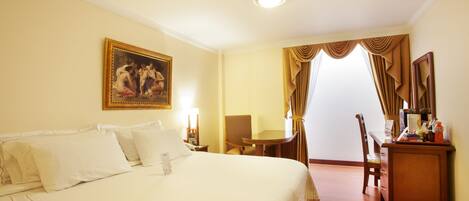 A hotel room with a large bed, a desk with a chair, a painting on the wall, and a view through a window with curtains.