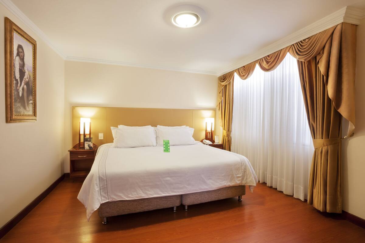 A hotel room with a large bed, two bedside lamps, a nightstand, a framed picture on the wall, and a window with curtains.