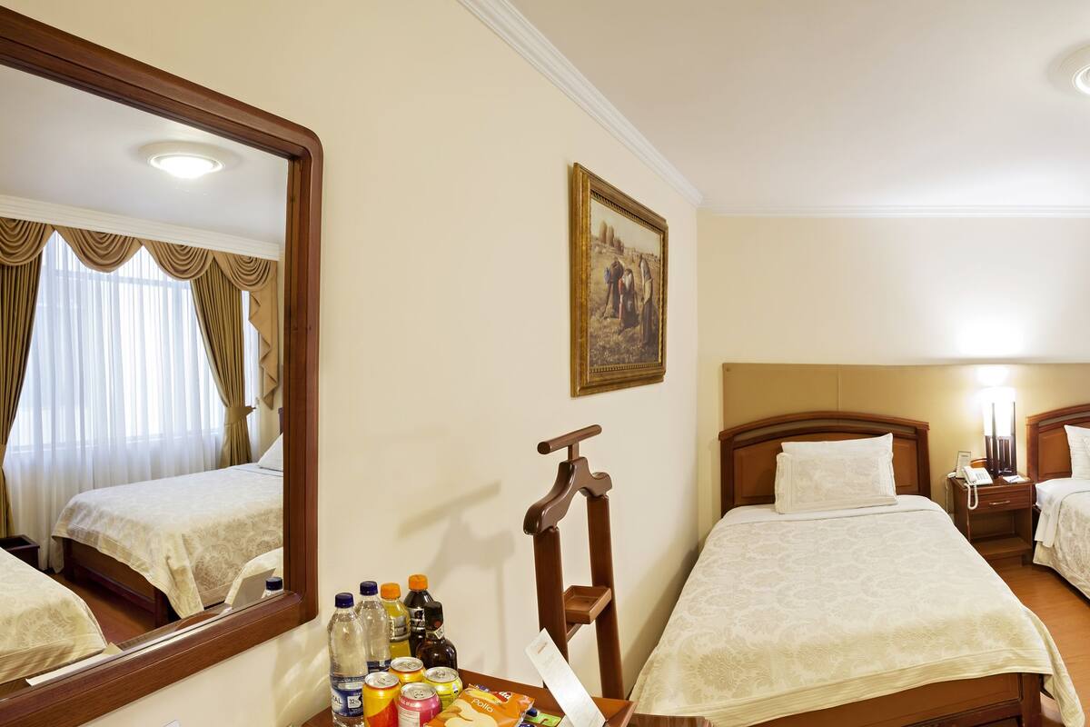 A hotel room with two single beds, a large mirror, a bedside table with a lamp, and a small table with snacks and drinks.